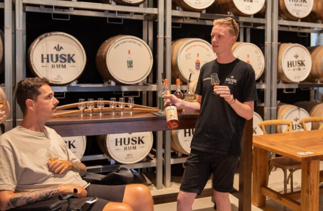 A man in a wheelchair enjoys a rum tasting during a distillery tour: he is holding a tasting glass and interacting with a guide, with distillery equipment visible in the background.